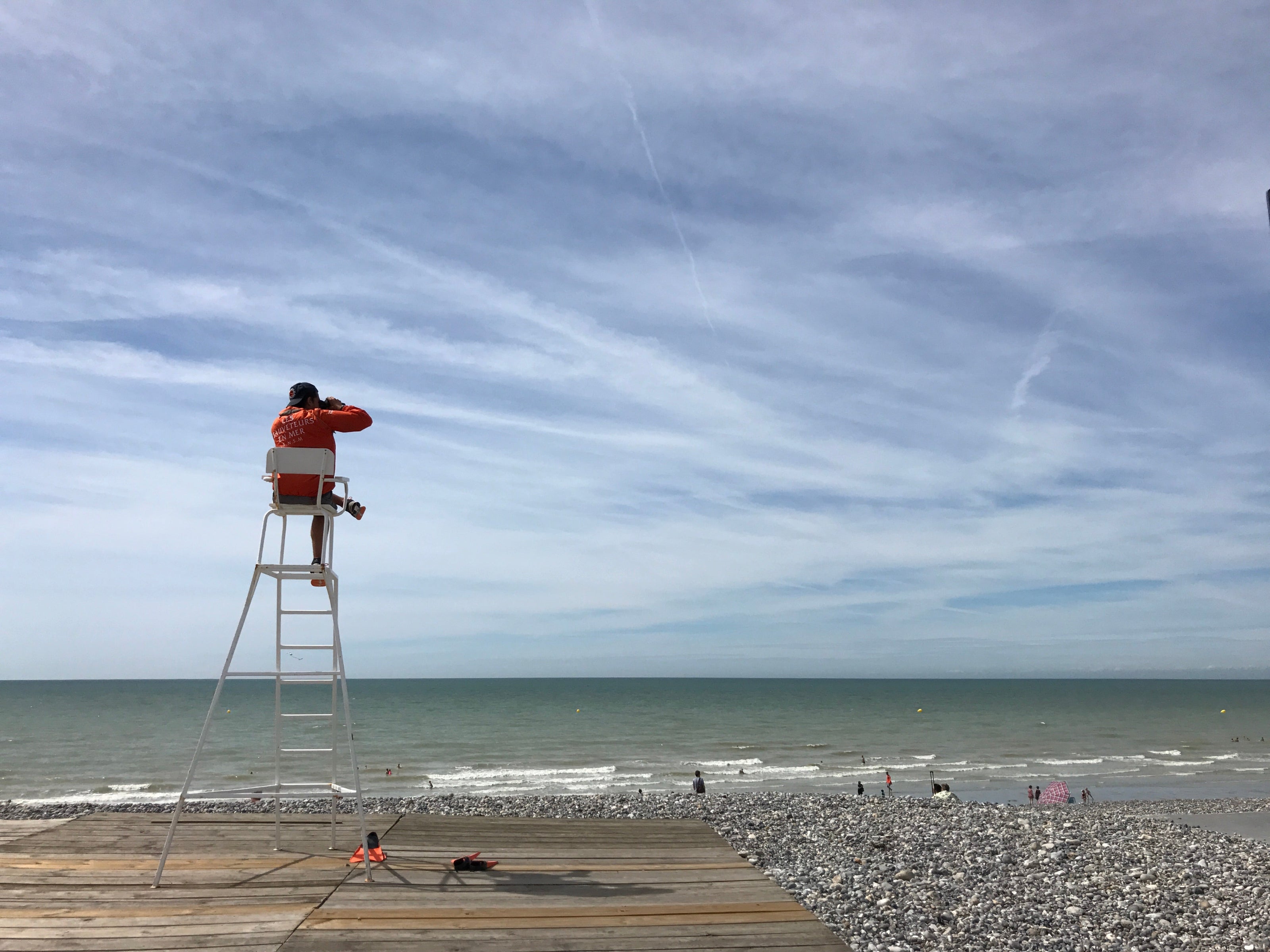 Person on a lifeguard tower overlooking the ocean under a blue sky with white clouds.